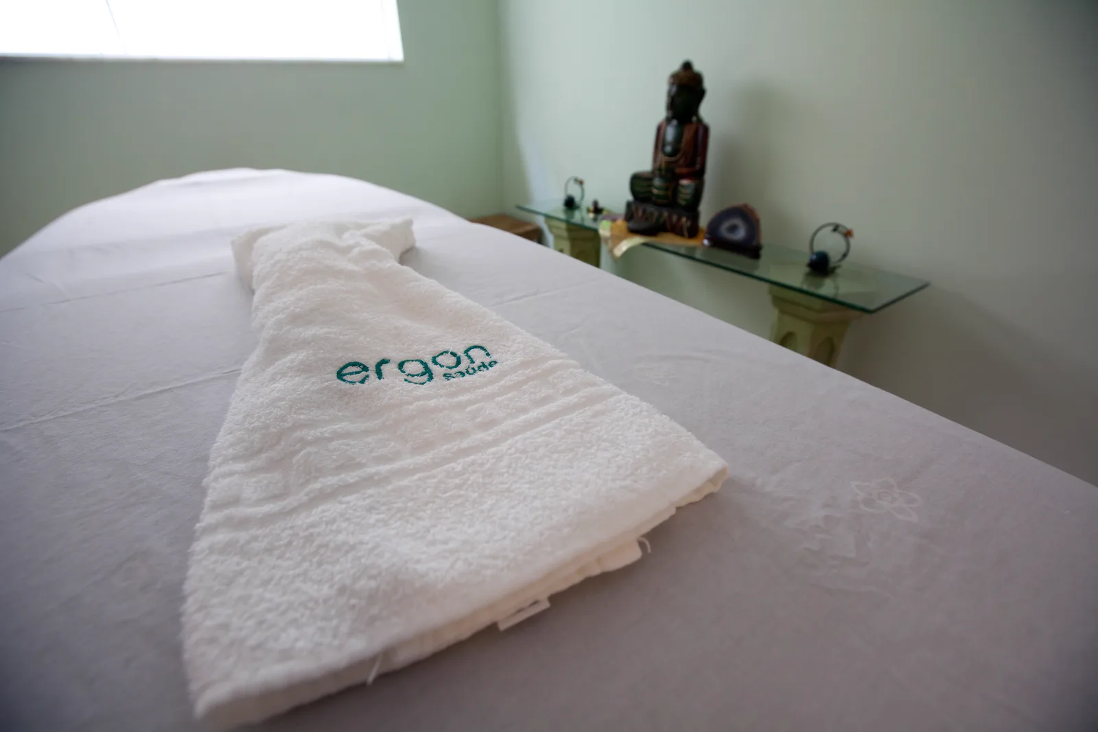 Toalha bordada Ergon Saúde sobre a maca de massagem, com buda ao fundo, na sala de massagem relaxante em Sorocaba